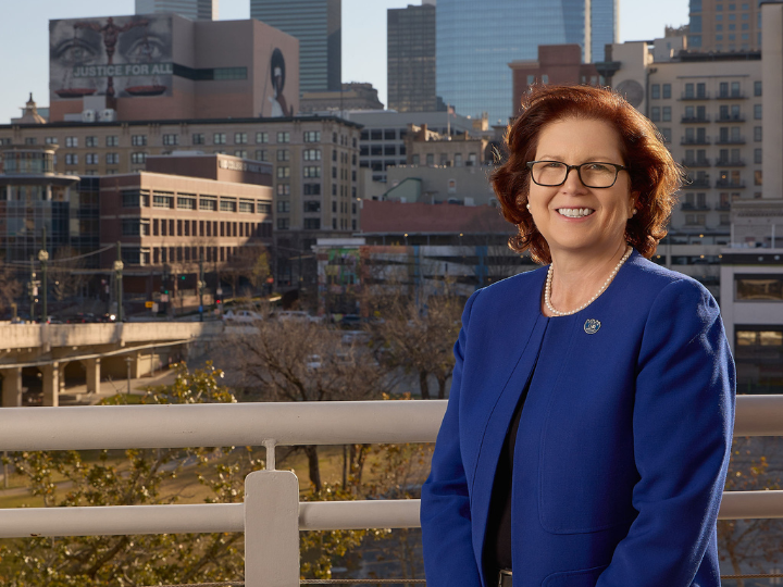 Smiling woman in blue jacket and pearl necklace posing in front of Houston’s city skyline.